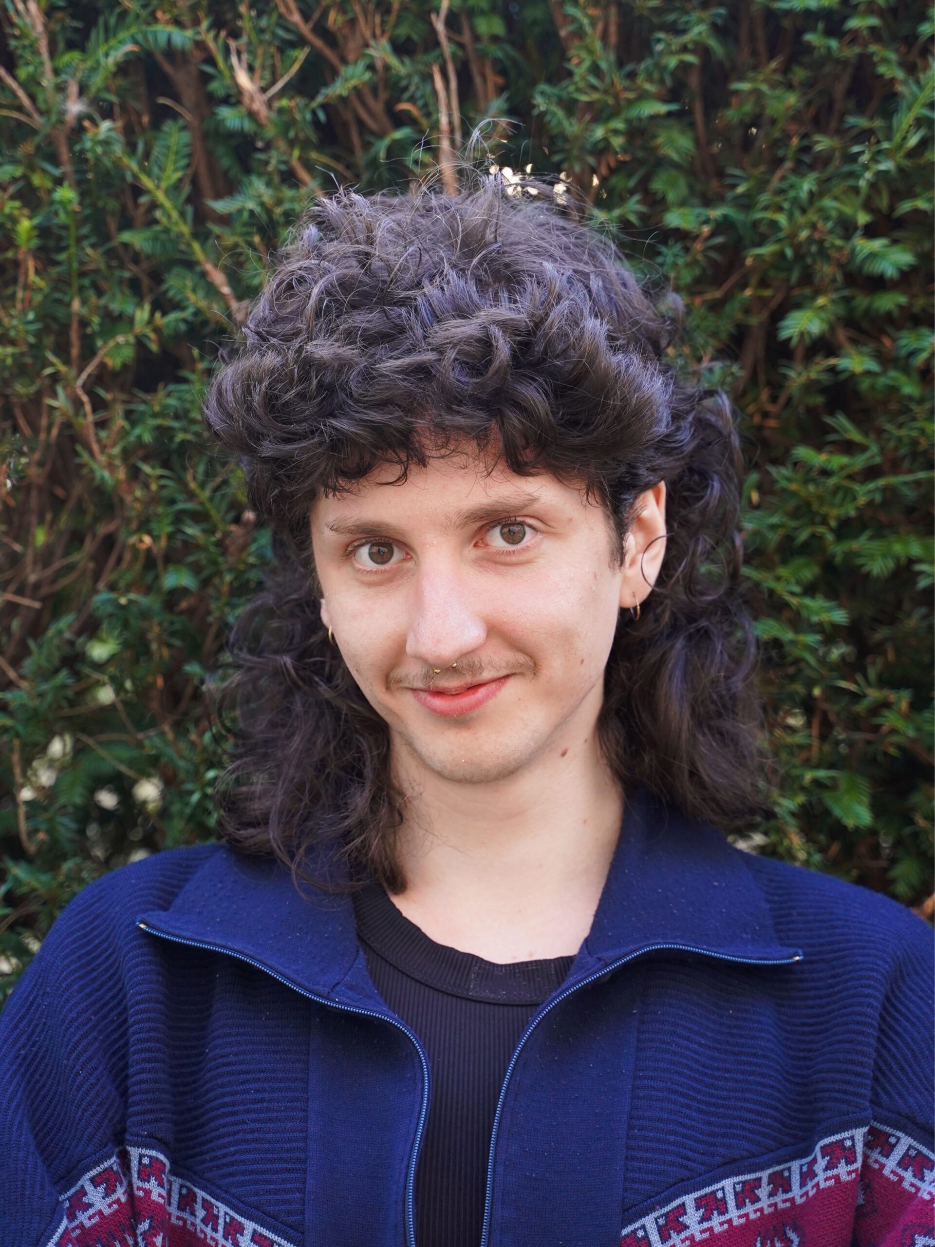 Eine junge Person mit lockigem, dunkelbraunem Haar posiert vor einer Hecke.