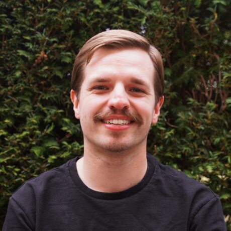 Ein junger Mann posiert vor einer Hecke. Er hat kurzes braunes Haar, trägt einen schwarzen Pullover und lächelt in die Kamera.