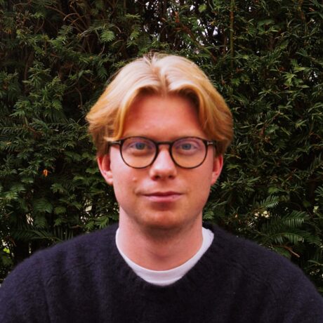 Porträtfoto eines jungen Mannes mit kurzem, blonden Haar und runder Brille. Er trägt einen dunkelblauen Pullover über einem weißen T-Shirt und blickt lächelnd in die Kamera. Im Hintergrund ist eine dichte grüne Hecke zu sehen.