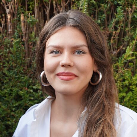 Eine junge Frau posiert vor einer grünen Hecke. Sie hat lange Braune Haare, trägt ein weißes Hemd und lächelt in die Kamera.