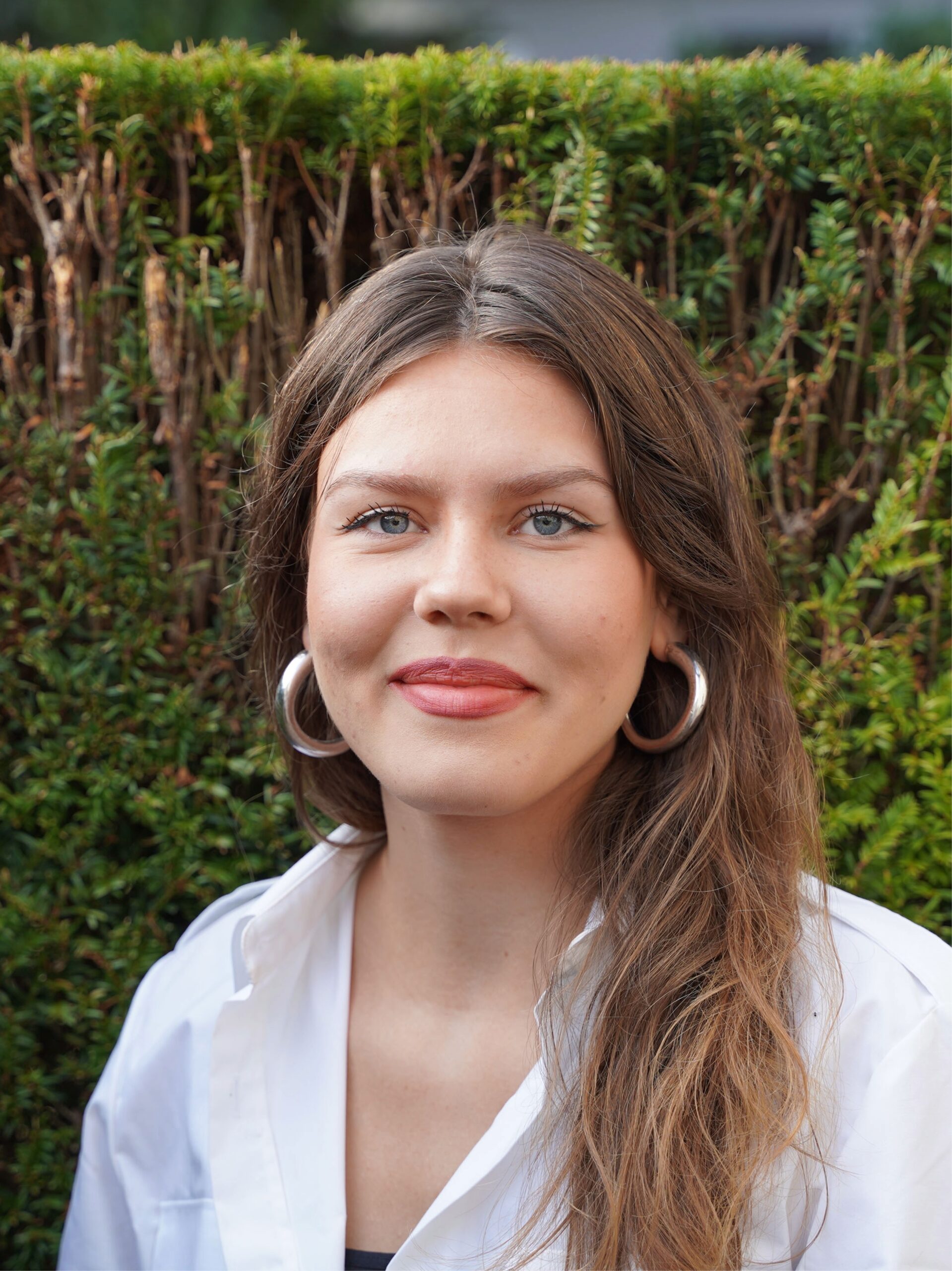 Eine junge Frau posiert vor einer grünen Hecke. Sie hat lange Braune Haare, trägt ein weißes Hemd und lächelt in die Kamera.