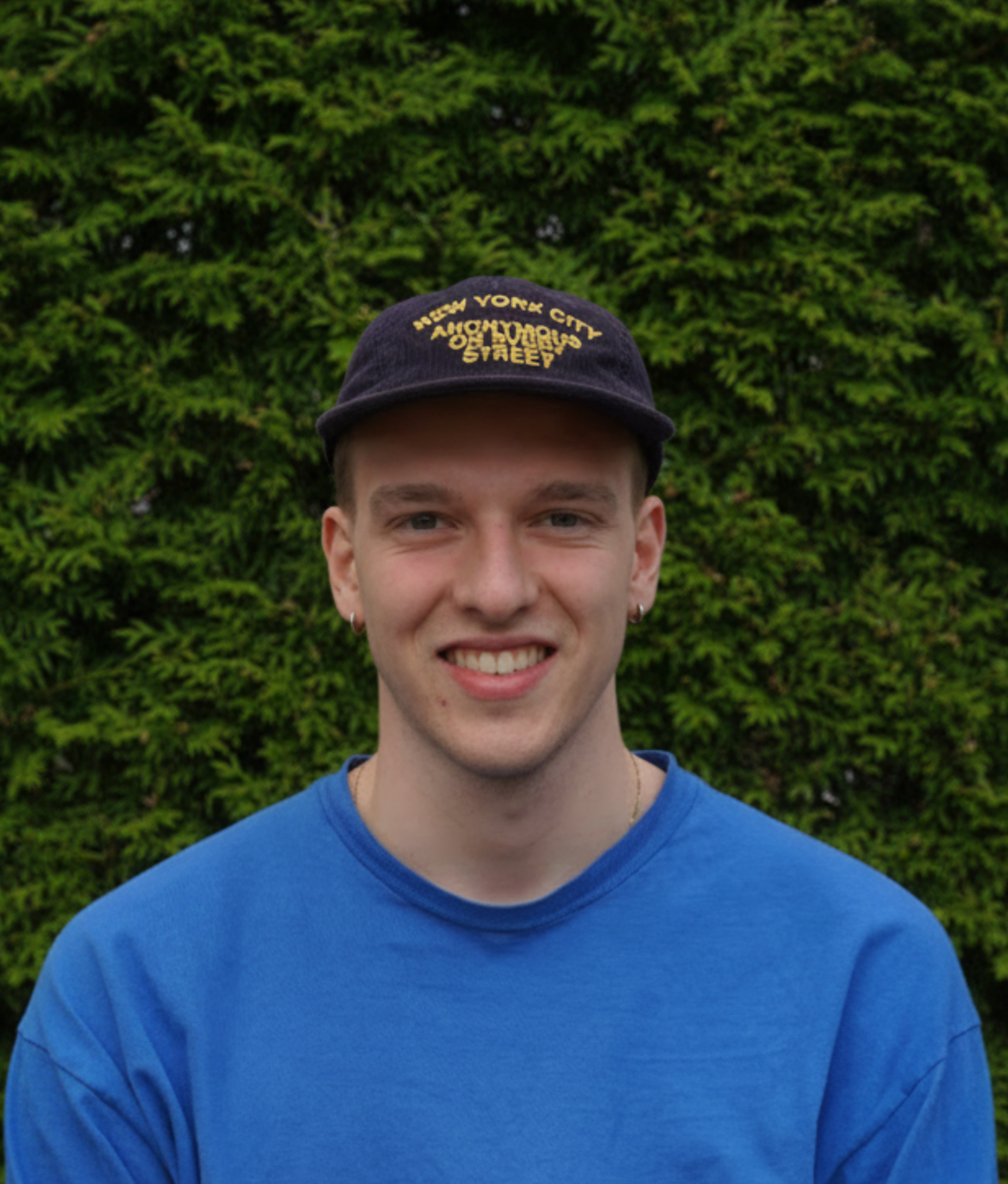 Ein junger Mann posiert vor einer Hecke. Er trägt eine dunkelblaue Mütze, einen blauen Pulli und lächelt in die Kamera.