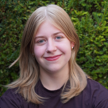 Eine junge Frau mit schulterlangem, blonden Haar und braunem Oberteil posiert vor einer Hecke. Sie lächelt in die Kamera.