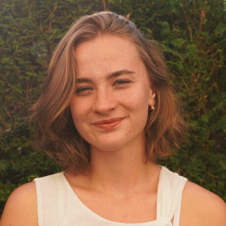 Porträt einer jungen Frau mit kinnlangem, blondem Haar. Sie trägt ein weißes Oberteil und blickt lächelnd in die Kamera. Im Hintergrund ist eine dichte grüne Hecke zu sehen.
