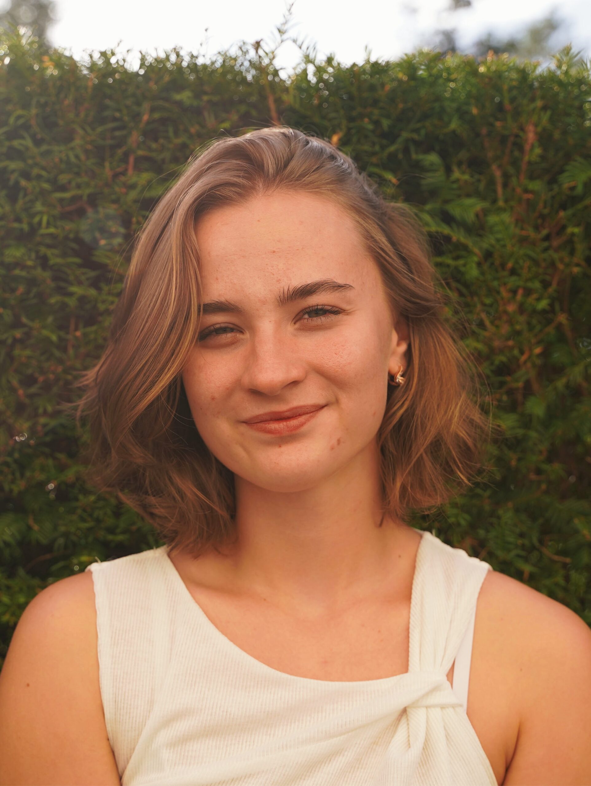 Porträt einer jungen Frau mit kinnlangem, blondem Haar. Sie trägt ein weißes Oberteil und blickt lächelnd in die Kamera. Im Hintergrund ist eine dichte grüne Hecke zu sehen.